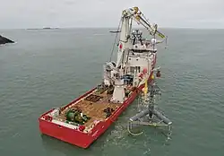 A workboat with a large crane is lowering a tidal turbine mounted on a triangular steel frame into the sea.