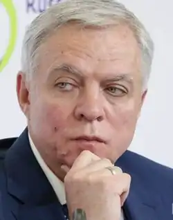 Grey-haired caucasian male wearing a suite and tie attending a press conference, hand on chin