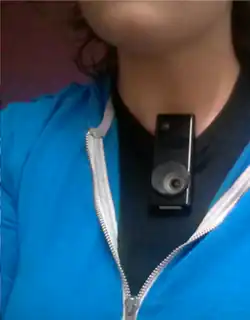 A close up of a person's neck and upper torso, with a black rectangular sensor and camera unit attached to their shirt collar