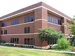 A three-story rectangular brick building seen from its corner. There is a small tree near its corner, and grass in the foreground.