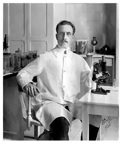 Black and white photo of Charlos Chagas, in his lab coat, sitting next to his microscope and surrounded by flasks and jars