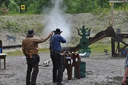 A Cowboy action shooter firing a lever action rifle at steel targets. The Range Officer to the left is holding a shooting timer to measure the time.