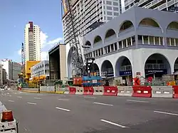 A part of the road has been blocked off with road barriers. Two cranes (one in the forefront) towers over the buildings beside the road.
