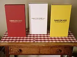 Three large game cases resembling books or binders sitting on a table, consisting of a maroon one labeled Famicom Mini, a white one labeled Famicom Mini Volume 2, and a yellow one labeled Famicom Mini Disk System Selection, all subtitled 20th Anniversary Family Computer.