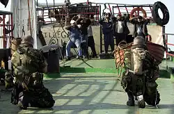 On a boat, two soldiers are pointing guns at eight Arabic men. The soldiers are in green camouflage, they're kneeling and only their backs can be seen. One of them is wearing a "camel water bag" on his back. The Arabic sailors are wearing civilian clothing and all of them have their hands on their heads.