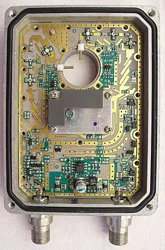 A low-noise block converter with the lid and horn removed exposing the complex circuitry inside, with the exception of the local oscillator which remains covered. The horizontal and vertical polarisation probes can be seen protruding into the circular space where the horn is normally attached. Two output connectors can be seen at the bottom of the device.