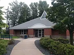 The Minnetonka Library, a one-story building with a large, triangular roof, out of which a triangular skylight arises. The building is situated among tall trees, and has a path leading away from its simple entrance. The path is adorned with bushes and a bench.