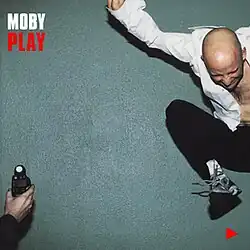 Album cover for Moby's "Play" featuring Moby jumping in an open white shirt in front of a teal wall, before a light meter