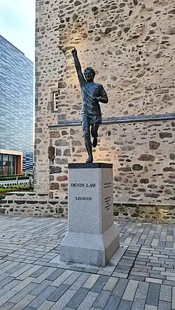 a bronze statue of a footballer, one arm raised, finger pointing to the sky, mounted on a granite plinth.