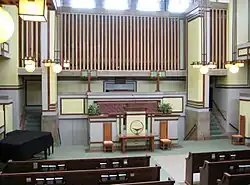 Interior of the auditorium as seen from the balcony. There is a wooden pulpit near the wall at the far end. Stairs ascend and descend on either side of the pulpit.