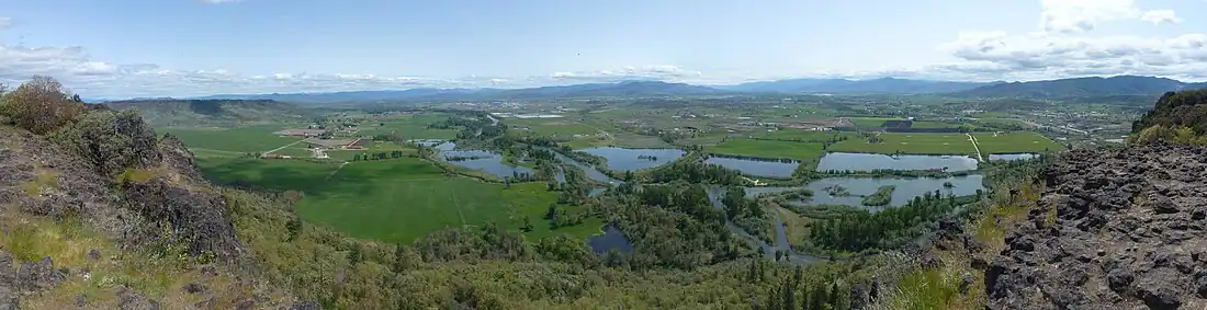 A river, seen from above, winds through flat farmland and a series of ponds on both sides of the stream. Buildings are scattered here and there across the landscape under a cloudy sky.