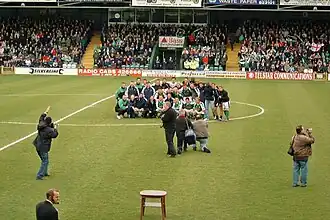 A view inside a football stadium. The winning team are posing together for a photograph, and there are photographers and journalists on the pitch.